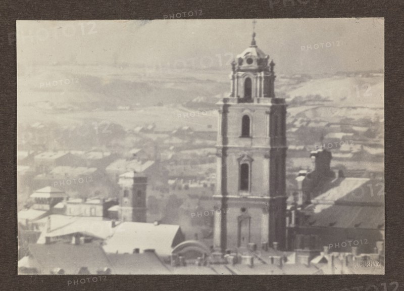 St. John's Church; photograph, 1914; subject: panoramic view of Vilnius including St. John's Church; photographer: Jan Bulhak 1876–1950; place of origin: Vilnius, Lithuania; medium: photographic print; image dimensions: 11.2 × 16.0 cm; album page dimensions: 31.0 × 23.5 cm., A grayscale photograph showing a tall, multi-tiered tower with arched openings and architectural detailing rising above a townscape; the tower is positioned toward the right side of the frame and is surrounded by a group of lower rooftops and building forms with sloped and flat surfaces. The foreground includes a cluster of roofs and chimneys, while the midground shows additional buildings and another smaller tower partially obscured. The background fades into distant, hilly terrain and soft atmospheric haze. Visible colors are shades of gray, off-white, and black.