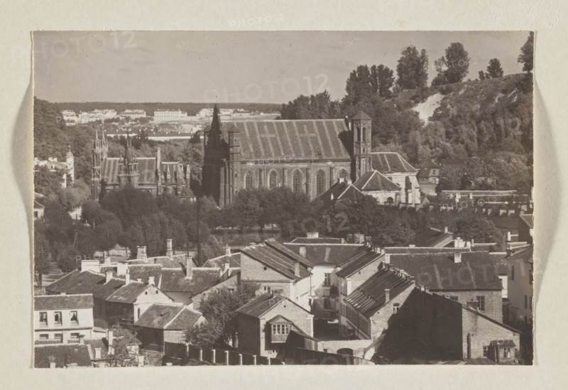 Bernardine Church; photograph, 1912, panoramic view of Vilnius; photographic print, height 10.5 cm, width 15.8 cm; album page height 31.0 cm, width 23.5 cm., The image shows a sepia-toned scene of a town with a group of rooftops in the foreground, composed of pitched roofs, chimneys, and brick and plaster facades in tones of brown and tan; behind the rooftops is a cluster of trees in darker browns and muted greens, and beyond the trees stands a large stone building with a steep roof and tall towers in darker brown tones; further back are rolling wooded hills with a mix of darker and lighter greenish-brown foliage and a lighter patch on a slope, and the sky above is a pale beige-gray.