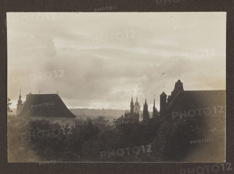 View from Užupis; photograph, Bulhak, Jan 1876–1950, panorama of Vilnius, 1912, Vilnius, photographic print: 10.6 × 15.4 cm; album page: 31.0 × 23.5 cm, The image shows a sepia-toned cityscape with a horizon of cloudy sky above a silhouette of buildings and trees, featuring a group of church spires and domes rising against the sky, dark rooflines and towers in the foreground, and a mass of treetops at the bottom; visible colors include shades of brown, beige, black, and gray.
