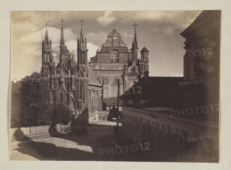 Church of St. Anne and the Bernardines; photograph, 1912. Subject: Bernardine Church Vilnius and St. Anne's Church Vilnius, Gothic style. Photographer: Jan Bulhak 1876–1950. Medium: photographic print; image size 11.6 x 15.8 cm; album page size 31.0 x 23.5 cm., A sepia-toned photograph showing a street flanked by architectural structures, with a large ornate Gothic-style church dominating the left and center featuring tall spires, pointed arches, and intricate stonework, and a long building with repeating arched details running along the right side and foreground. A few trees and foliage appear near the church, and a low picket fence lines part of the street. Shadows stretch across a cobbled or textured roadway where a small cart or wagon and a couple of indistinct figures are visible. The sky shows a few clouds above the buildings. The visible colors are shades of brown, tan, beige, and muted gray.