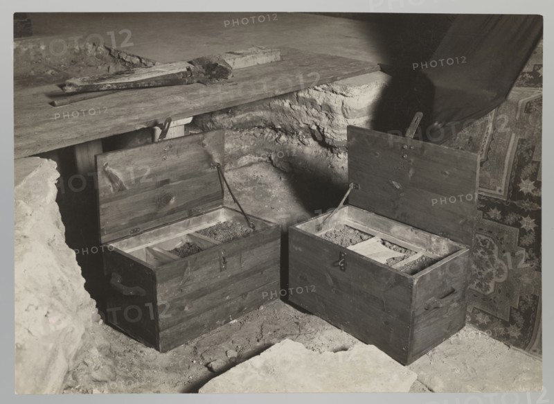 Royal Cathedral Crypt — Open Chests; Bulhak, Jan 1876–1950; Vilnius Lithuania; Archcathedral Basilica of St. Stanislaus and St. Ladislaus; crypts; 1931 flood in Vilnius; 1931; photograph; nature photograph; height 12.0 cm; width 16.0 cm, The image shows a grayscale scene of an indoor area with a rough stone or dirt floor and a wooden platform; two wooden chests with open lids sit side by side on the floor, each chest divided into compartments containing loose granular material; the chests have dark wood tones and metal hinges and handles, and there are a few tools or wooden pieces resting on the platform above; a patterned textile with intricate designs hangs or is draped on the right side, showing darker and lighter areas; shadows fall across the scene, creating contrasts between light and dark gray tones.