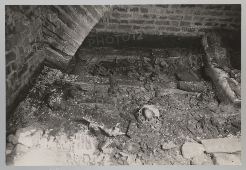 Vilnius. Cathedral — interior of the royal crypt. Photograph from nature, 1931. Depicts crypts and archaeological excavations following the 1931 flood in Vilnius. Photographer: Jan Bulhak 1876–1950. Place: Vilnius Lithuania, Archcathedral Basilica of St. Stanislaus and St. Ladislaus. Photograph; 11.5 × 16.5 cm., The image shows a dim interior space with brick walls and an arched brick structure, a rough dirt and debris-covered floor, and scattered wooden beams and broken planks. There are several irregular chunks of rubble and fragments of material spread across the ground, along with a partially exposed rounded object among the debris. The visible colors are shades of gray, black, and white.