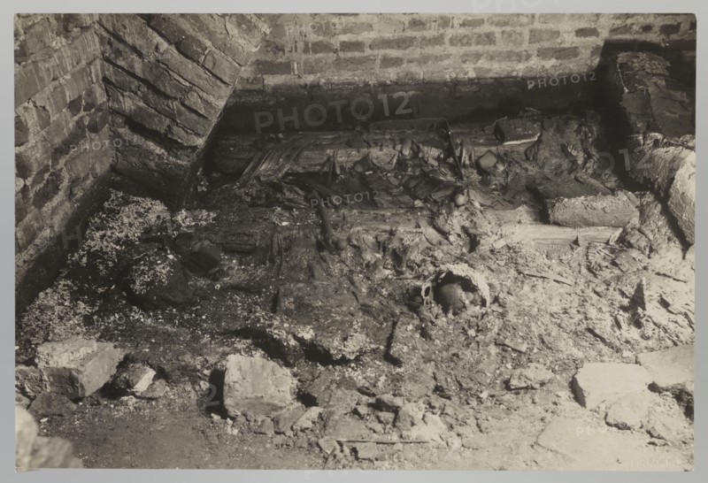 Vilnius. Cathedral - interior of the royal crypt, Bulhak, Jan 1876-1950, Vilnius Lithuania, Archcathedral Basilica of St. Stanislaus and St. Ladislaus, crypts, archaeological excavations, flood in Vilnius in 1931, 1931, photograph, photograph from nature, height 11.5 cm, width 16.5 cm, A close-up view of a rough, enclosed area with brick walls and a dirt floor showing debris and broken materials; the scene includes a brick corner with angled mortar lines, scattered chunks of masonry and rubble, splintered wooden planks and fragments, and areas of crumbly, uneven ground with dust and small granular particles. The visible colors are shades of black, white, and gray.