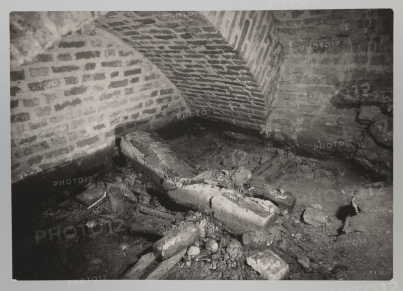 Vilnius. Cathedral — interior of the royal crypt, photographed by Jan Bulhak 1876–1950. Archcathedral Basilica of St. Stanislaus and St. Ladislaus, crypts; archaeological excavations; associated with Barbara Radziwillówna Queen of Poland, 1520–1551. Flood in Vilnius, 1931. Photograph from nature; gelatin silver print, — height 11.5 cm, width 16.5 cm., The image shows a dimly lit interior space with brick walls and a curved brick ceiling; the bricks display various shades of gray. In the foreground there is a damaged, broken rectangular stone or concrete object surrounded by rubble and fragments, all in tones of gray and black. The floor appears uneven and dirty with scattered chunks of masonry and loose debris in darker and lighter grays. Shadows create contrast along the base of the walls and among the rubble, producing areas of deep black and mid-gray. Overall the visible colors are shades of gray, black, and white.
