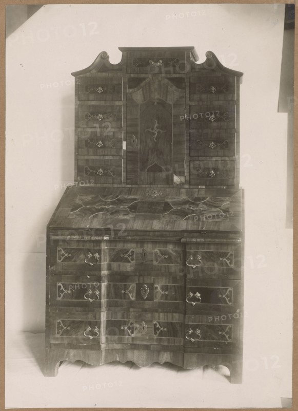 Secretary with setting from the mid-18th century — general view; maker: Józef Kosciesza Jaworski fl. ca. 1911–ca. 1929 photographer; furniture, baroque style; ; intarsia, artistic crafts; bureaus/desks; 18th-century craftsmanship; lost monuments partially; print with entire surface re-glued onto cardboard; not after 1925; places of origin: Kraków original, Warsaw; photography/reproduction of a work of art; photographic print: height 22.0 cm, width 15.8 cm; cardboard: height 34.8 cm, width 25.0 cm., The image shows a tall wooden piece of furniture with a darker wood grain and decorative inlay, consisting of an upper cabinet section with an ornate curved top and multiple small drawers flanking a central door, and a lower section with a slanted writing surface above a set of larger drawers; the hardware includes several metallic handles and escutcheons, and the piece rests on a scalloped base. The visible colors are shades of brown in the wood, metallic gray on the hardware, and pale off-...