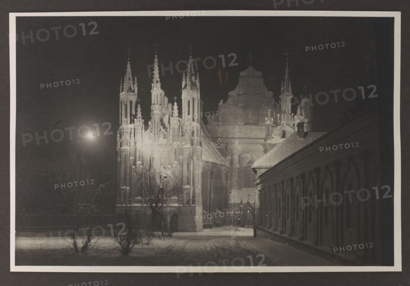 Vilnius Illuminated. Churches of St. Anne and the Bernardines; Edmund and Boleslaw Zdanowski, photographers; Vilnius Lithuania, Bernardine Church, St. Anne's Church; before 1939; photograph, photographic print: height 8.5 cm, width 13.5 cm; album page: height 17.0 cm, width 24.5 cm, A nighttime scene showing a large gothic-style building with tall spires and ornate architectural details illuminated against a dark sky, a smaller building with a sloped roof and a row of arched openings extending along the right side, a snow-covered foreground with a few leafless trees and low shrubs, a bright light source in the sky casting light across the scene, and visible tones of black, dark gray, light gray, and white.