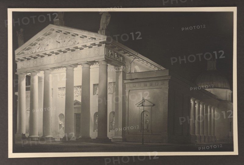 Vilnius Illuminated. Cathedral, facade and southern elevation, photographed by Edmund and Boleslawa Zdanowscy; Vilnius Lithuania; cathedral Vilnius; classicism architect; before 1939; photograph, photographic print: height 8.5 cm, width 13.5 cm; album page: height 17.0 cm, width 24.5 cm, A nighttime scene of a classical building facade with a row of large, round columns supporting a triangular pediment; the facade and columns are lit in pale tones while the background is dark, creating strong contrasts of light and shadow. The pediment and entablature show carved reliefs and decorative details. Between the columns are recessed niches each containing sculptural figures, and there is a taller rectangular section to the right with a recessed statue in a framed niche and a slender street lamp in front. To the far right a colonnaded extension with several columns leads toward a domed structure partially visible behind it. The image shows shades of black, gray, cream, and sepia.