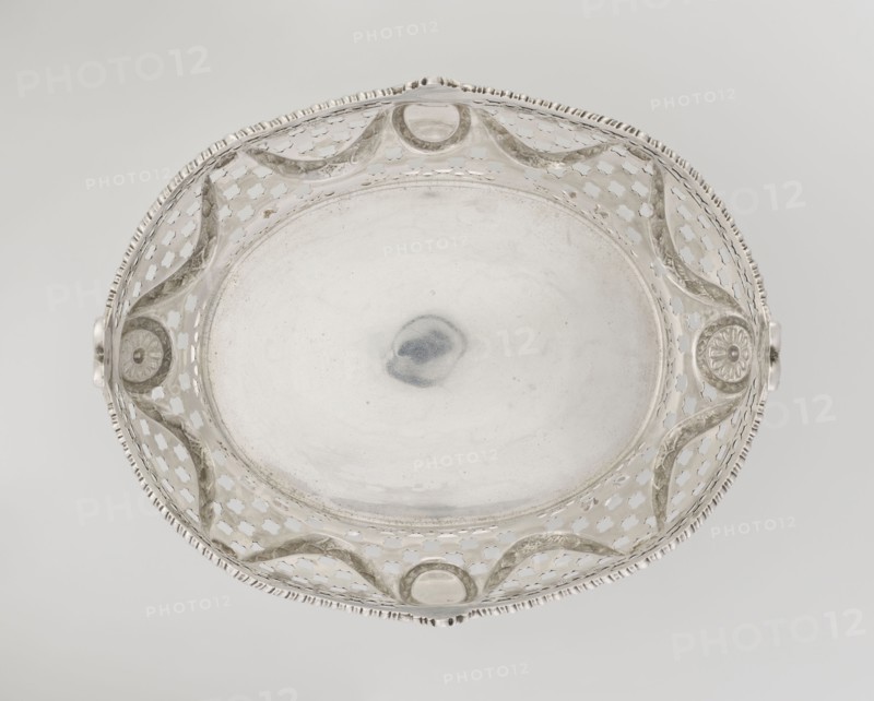 Openwork fruit basket by Harris, CS & Sons London; goldsmith's firm; fl. 1817–1933 and Charles Stuart Harris goldsmith; fl. 1852–1897; inscription/association with Ignacy Jan Paderewski 1860–1941. Oval openwork fruit basket supported on four palmette legs with the lower edge decorated with U-sections; sides feature openwork circle and trellis motifs with applied laurel garlands, medallions and rosettes; wavy-cut upper edge decorated with a godron ribbon, raised on the longer axis and decorated with acanthus leaves, on the shorter axis with palmettes. Materials: presumably silver; decorative elements include palmettes, bay leaves, garlands, godron ornaments, medallions, rosettes. Date: 1896/1897. Place: London, England, Great Britain. Function: vessel fruit basket. Dimensions: height 11.2 cm; width 23 cm; depth 18.5 cm; weight 645 g., A round metallic dish photographed from above with a central smooth shallow basin surrounded by an openwork rim featuring repeating cutout shapes and scal...