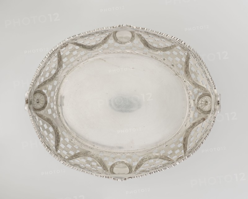 Openwork fruit basket by Harris, CS & Sons London; goldsmiths, fl. 1817–1933 and Charles Stuart Harris fl. 1852–1897. Oval openwork fruit basket supported on four palmette legs with a lower edge decorated with C-sections. Sides feature openwork composed of circle and grid motifs with applied laurel garlands, medallions, and rosettes. Upper edge cut in a wavy pattern and decorated with a godron ribbon; raised on the longer axis and decorated with acanthus leaves, and on the shorter axis with palmettes. Contains palmettes, bay leaves, garlands, godron ornaments, and medallions., A shallow round decorative dish viewed from above with an openwork rim featuring repeating quatrefoil-shaped cutouts and undulating raised bands; the central flat surface is smooth and reflective. The rim has a notched edge and two small protruding handles with floral rosette motifs where they attach to the rim. The object appears metallic with a predominantly silver color and subtle variations including slightly...