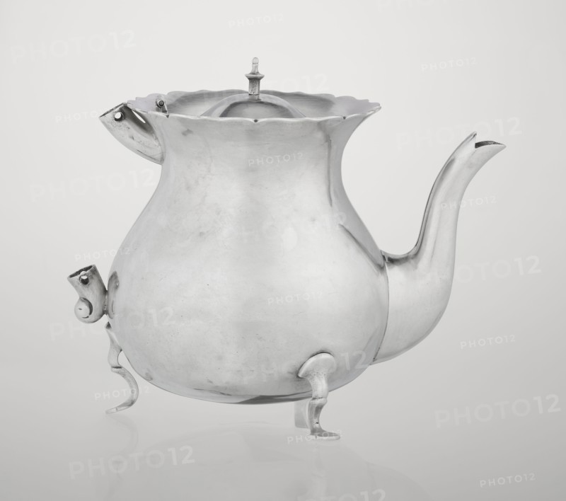Pear-shaped teapot supported on three legs, smooth, with a flared upper edge topped with a pinnacle, with a spout-shaped spout, with silver bushings on the axis for mounting a missing wooden handle. Maker: Stokes & Ireland Ltd Birmingham; goldsmiths; fl. 1872–1925. Date: 1900–1901. Place of origin: Chester, England, Great Britain. Materials/function: tea vessel. Dimensions: height 11.5 cm; width 15.5 cm; depth 10 cm; weight 330 g., A metallic teapot with a bulbous body and a wavy rim stands on a reflective surface, showing a smooth, slightly worn silver finish with subtle scuffs and darker tarnish marks; it has a curved spout extending outward and upward, a hinged lid with a small finial on top, a curved handle-like projection opposite the spout, and three short ornate legs supporting the body, all in silver tones against a pale gray background.