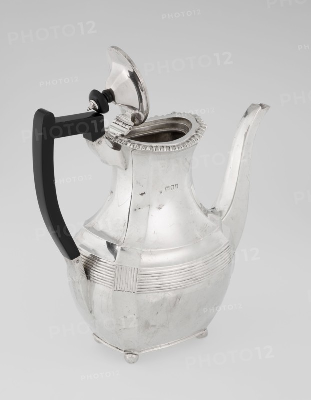Coffee pot by Harris, CS & Sons London; goldsmiths' firm, fl. 1817–1933 and Charles Stuart Harris maker, fl. 1852–1897. Purchased; scrap — purchased. Pear-shaped, ribbed coffee vessel with godron ornaments; belly decorated with a band of narrow half-cylinders and grooves. Supported on four ball-shaped, profiled, ribbed legs. High side spout-shaped handle and a contemporarily added wooden axial handle set in silver sockets. Upper edge edged with godron band; hinged lid domed in the center, topped with a bowling-pin finial. Dated 1895/1896. Great Britain, London, England. Height 21.2 cm; width 18.5 cm; depth 10 cm; weight 640 g., A metal pitcher with a hinged lid and tall curved spout sits against a pale background, the pitcher showing a reflective silver surface with visible scratches and small dents; it has a rounded body that tapers to a narrower neck with a decorative ridged rim and a band of horizontal grooves around the midsection interrupted by a textured rectangular panel, and it...