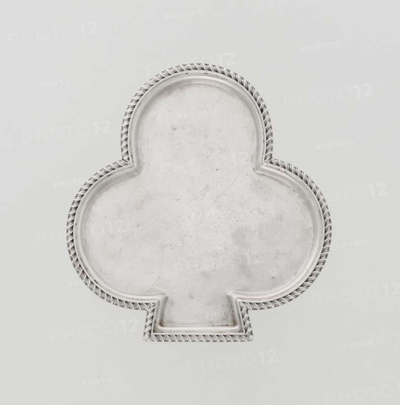 Tray, club-shaped with a smooth mirror surrounded by a raised half-cylinder edged with diagonal godron ornament. Maker: Hukin & Heath Birmingham; London; goldsmiths' firm; fl. 1855–1953; makers: John Thomas Heath and John Hartshorne Middleton. Date: 1897–1898. Place: London, England, Great Britain. Materials/subject: table accessory. Dimensions: height 1.1 cm; width 12.5 cm; depth 11.9 cm; weight 86 g. Provenance: purchase; scrap — purchase., A metallic tray shaped like a club with a raised decorative twisted rope border, showing a worn and slightly scratched surface; colors visible include silver, gray, off-white, and a pale background with subtle shadows.