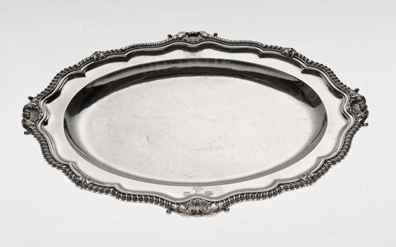 Oval platter by Benjamin & James Smith fl. 1809–1812, goldsmiths; Georgian/George III style, oval vessel with smooth mirror and multi-leaf ribbed collar edged with a godron band with four shells on axes, enclosed within acanthus leaves; godron ornaments, shells, acanthus ornament. Engraved coats of arms on collar; underside of collar engraved monogram GvR, dated 1811/1812. Materials: goldsmith's work. Dimensions: height 3.5 cm; width 58.5 cm; depth 45.4 cm; weight 3550 g. Places: Great Britain, England, London., A large oval metal tray with an ornate scalloped rim and decorative handles rests against a pale background; the tray has a reflective silvery surface with visible fine scratches and wear, and the rim features detailed embossed patterns and flowing curves in shades of silver and gray.
