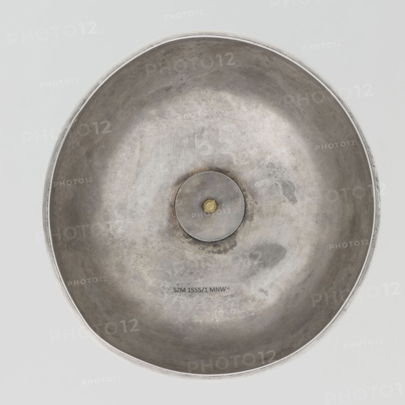 Platter lampshade, Scott, Digby & Smith, Benjamin fl. 1802–1807 goldsmith; silver hemispherical lampshade with a non-original handle, smooth, ribbon edge, profiled. Relief plaque with the Czartoryski coat of arms on a paludament, under a princely mitre, with labres and the motto Be that as it may. Handle is contemporary. 1803/1804, London, England, Great Britain. Vessel; height 8.5 cm, diameter 15.2 cm, weight approx. 230 g., A large round metallic bowl seen from above with a shallow concave interior showing a weathered, uneven surface in shades of silver and gray and a few darker gray patches, a circular metal disc with a small golden element at its center is visible in the middle, the rim is a slightly brighter silver and the background around the bowl is a pale gray.