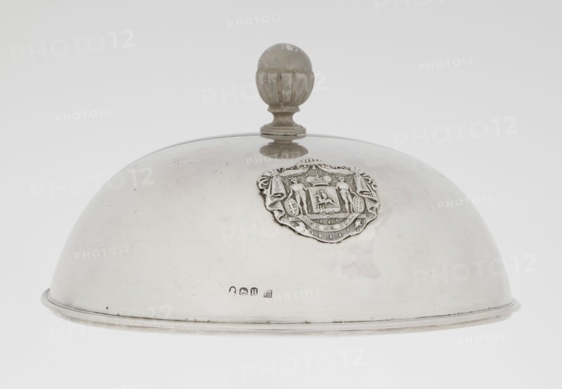 Platter lampshade by Scott, Digby & Smith, Benjamin fl. 1802–1807, goldsmith. Silver hemispherical lampshade with a non-original handle, smooth with a ribbon profiled edge. On the side, a relief plaque with the Czartoryski coat of arms on a paludament, under a princely mitre, with labres and the motto Be that as it may. The handle is contemporary. Dated 1803/1804, London, England, Great Britain. Vessel; height 8.5 cm; diameter 15.2 cm; weight approx. 230 g., A domed metal cloche with a round finial handle on top sits against a plain light background; the cloche is smooth and reflective with visible scuffs and scratches and a worn metallic sheen in silver and gray tones, and a decorative raised emblem with intricate sculpted details is attached to one side showing textured relief in similar silver-gray metal, while the finial appears slightly darker with muted gray and brownish discoloration, and the rim shows a thin darker band of metal.