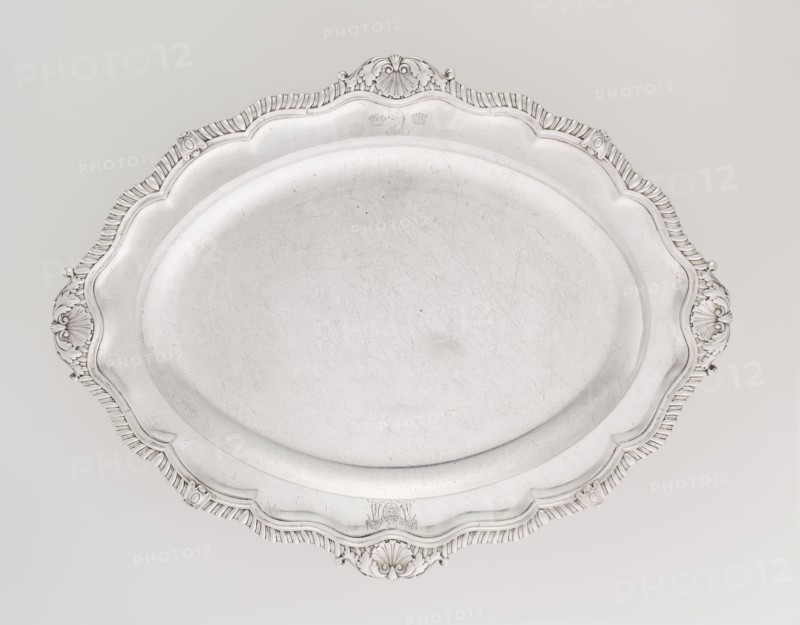 Oval Platter, Smith, Benjamin & James fl. 1809–1812 maker's role / goldsmith's firm; oval silver vessel, Georgian/George III style; smooth mirror with multi-leafed ribbed collar edged by a godron band with four shells on the axes, enclosed within acanthus leaves; engraved coats of arms on the shorter axis of the collar; underside of collar engraved monogram GvR, dated 1811/1812; England, London; height 3 cm, width 47 cm, depth 37.5 cm, weight 2420 g., A large oval metal tray with a smooth, slightly scratched central surface and a decorative scalloped rim featuring ornate shell and scroll motifs; the tray and its details appear in shades of silver and light gray against a pale off-white background.