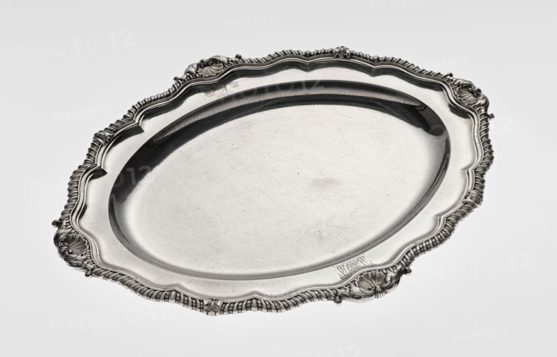 Oval platter by Benjamin & James Smith fl. 1809–1812, maker's mark; Georgian/George III style; silver vessel, oval, with smooth mirror and multi-leafed ribbed collar edged with a godron band with four shells on the axes, enclosed within acanthus leaves; engraved coats of arms on the shorter axis of the collar; underside of the collar engraved monogram GvR, dated 1811/1812; ornament: shells, godrons, acanthus leaves; dimensions: height 3 cm, width 47 cm, depth 37.5 cm; weight 2420 g; London, England, Great Britain., A large oval metal platter with an ornate raised rim featuring shell-like and rope-like decorative motifs, visible scrollwork, and embossed floral accents; the central surface is smooth with visible fine scratches and a slightly reflective finish. The colors visible are silver, gray, and hints of darker gray in the shadows and detailing.