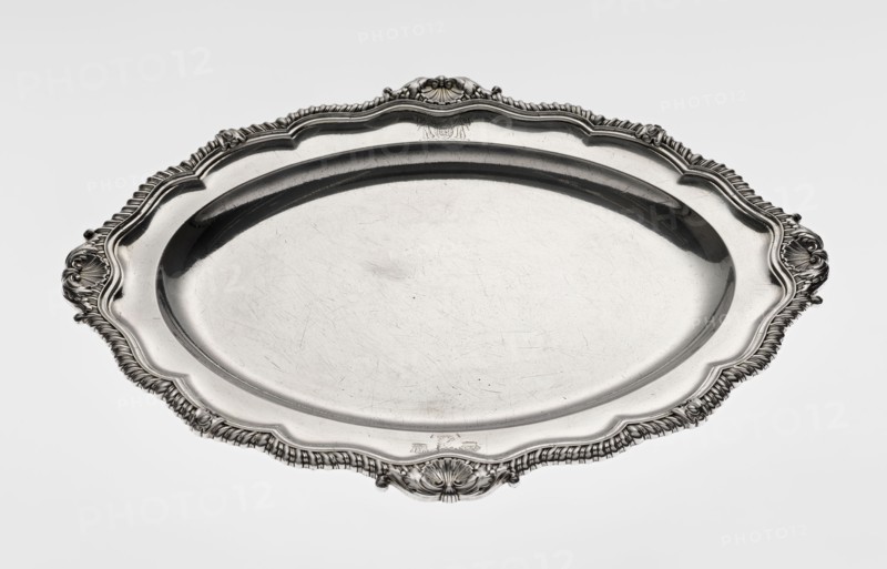 Oval platter by Benjamin & James Smith fl. 1809–1812, maker's mark; Georgian/George III style; silver; oval with smooth mirror and multi-leafed ribbed collar edged with a godron band and four shells on the axes, enclosed within acanthus leaves; engraved coats of arms on the shorter axis of the collar; underside of the collar engraved monogram GvR, dated 1811/1812; engraved shells, godron ornaments, acanthus leaves; vessel; height 3 cm, width 47 cm, depth 37.5 cm, weight 2420 g., A large ornate oval metal tray with a polished surface, showing a few fine scratches and scuffs, surrounded by an intricately molded rim with a scalloped edge and decorative shell- and scroll-like motifs at intervals; the tray appears reflective and metallic silver in color, with darker silver shadows in the recessed areas and along the inner rim, and lighter silver highlights on the raised decorative elements.
