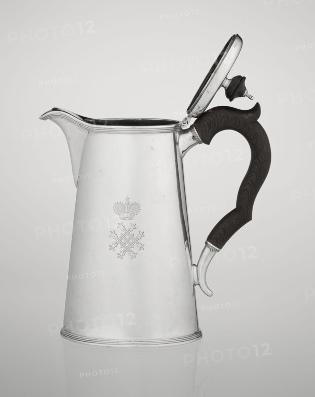 Conical coffee pot with hinged lid, slightly arched and flattened at the top, topped with a wooden handle; short spout on the axis with two sleeves for attaching a wooden handle added contemporarily. Engraved monogram tied HH under a ducal mitre; inscribed 1890/1891. Maker: Barnard, Edward & Sons Ltd London; goldsmith's firm; fl. ca. 1680– . Also associated maker/seller: Clark, Alfred Ltd London; goldsmith's firm and sellers; fl. 1898–1934. Materials: silver with wooden handle. Function: coffee ware. Place: Great Britain — England, London. Dimensions: height 17.3 cm; width 11 cm; depth 8.4 cm; weight 627 g., A metallic pitcher with a reflective silver surface and a hinged lid is shown against a light gray background, the lid open and revealing a dark interior; the pitcher has a curved spout and a dark brown wooden handle with visible grain and a carved contour, attached to the body with silver fittings, and there is an engraved decorative emblem on the pitcher’s front; the overall colo...