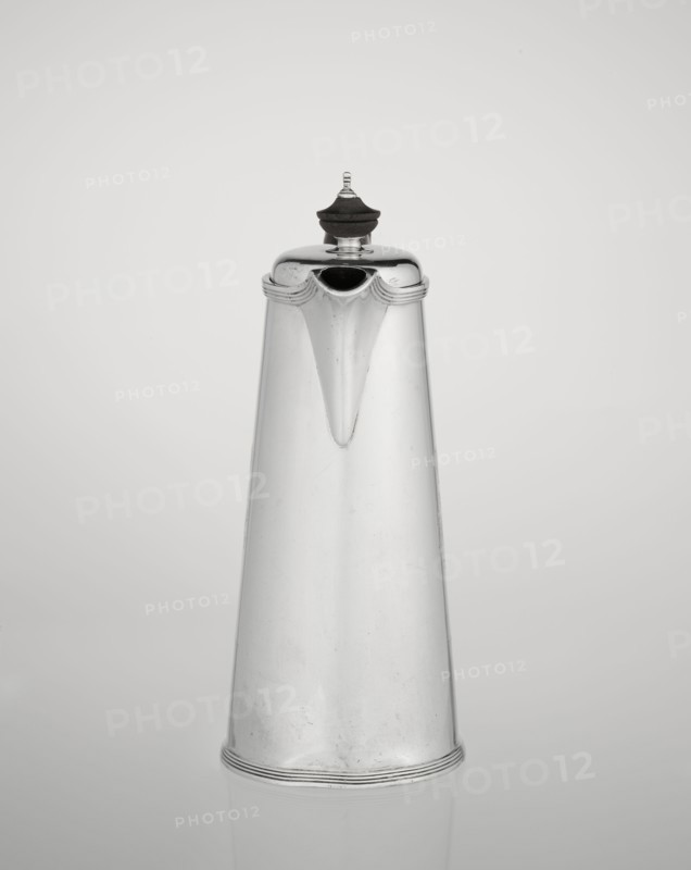Coffee pot by Barnard, Edward & Sons Ltd London; goldsmith's firm; fl. ca. 1680–, with later association to Clark, Alfred Ltd London; goldsmith's firm and sellers; fl. 1898–1934. Conical coffee pot with hinged lid, slightly arched and flattened at the top, topped with a wooden handle; short spout; on the axis two sleeves for attaching a wooden handle added contemporarily. On the mantle engraved monogram tied HH beneath a ducal mitre; dated 1890/1891. Material: silver with wooden handle. Place: London, England, Great Britain. Function: coffee ware. Dimensions: height 17.3 cm, width 11 cm, depth 8.4 cm; weight 627 g., The image shows a tall, tapered metal pitcher with a rounded lid topped by a dark finial; the pitcher has a smooth, reflective surface with subtle scratches and a small spout near the top, and it sits against a plain light gray background with a faint reflection beneath. The visible colors are silver, dark brown or black on the finial, and shades of light gray in the backgr...