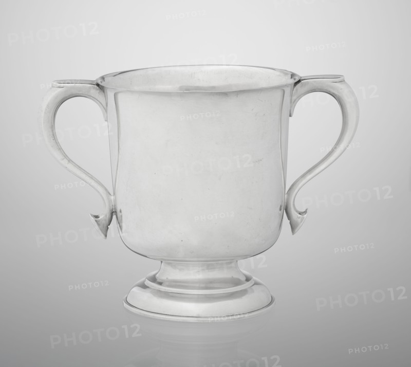 Two-eared vase, made by Hutton & Sons Ltd goldsmith and silversmith firm; fl. 1800–1930. Vase on a circular, stepped-profile foot and short stem; smooth cylindrical body with slightly upturned upper edge; two profiled volute-shaped handles on the sides. Dated 1926/1927. Great Britain — Birmingham, England. Vessel; height 15 cm, width 21 cm, depth 13.5 cm, weight 540 g., A plain metal cup with two curved handles sits centered against a smooth background; the cup has a rounded body that tapers to a stepped circular base and shows a reflective surface with slight scratches and subtle highlights, and the visible colors are shades of silver, gray, and white.