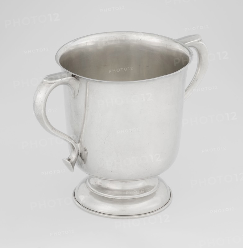 Two-eared vase, Hutton & Sons Ltd goldsmith and silversmith firm; fl. 1800–1930 maker, scrap - purchase, purchase provenance; vase on a circular stepped-profile foot and short stem, smooth cylindrical body with a slightly upturned upper edge, two profiled volute-shaped handles on the sides; 1926/1927; Great Britain, Birmingham, England; vessel; height 15 cm, width 21 cm, depth 13.5 cm, weight 540 g, A silver metal cup with a rounded body and a footed base sits against a light gray background; the cup has a smooth interior visible from above and two curved handles attached on opposite sides, each handle featuring a subtle decorative curve and a small pointed finial where it meets the body; the metal surface shows gentle reflections and a few faint scratches, and the overall colors visible are silver, gray, and the pale gray of the background.