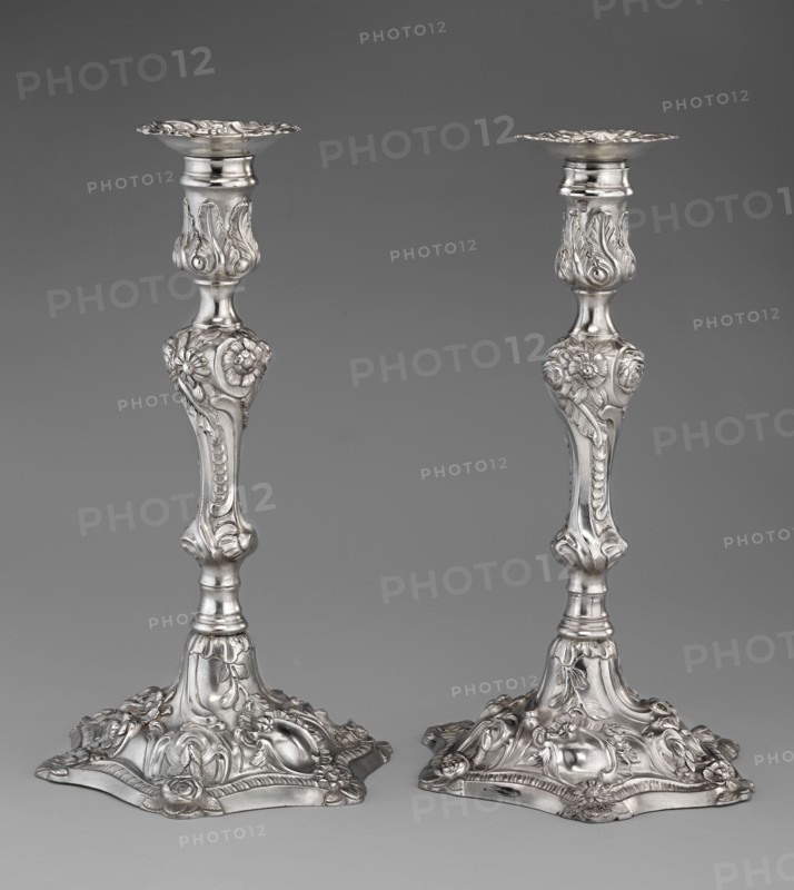 Single-candle candlestick by William Cafe fl. 1757–ca. 1802, rococo; silver. Rocaille ornament; plant ornaments, flowers, shells, cartouches, acanthus leaves; relief decoration. Rococo on a raised, domed foot with a hexagonal rim, decorated with flower heads at the corners, passing into a baluster shaft, terminating in a vase-shaped socket with a disc-shaped drip pan. Decoration slightly spirally twisted in George II / Georgian style. Dated 1763/1764. Place of origin: London, England, Great Britain. Function: lighting. Height 29 cm; diameter 16.9 cm; weight 695 g., A pair of ornate metallic candlesticks photographed against a plain gray background, each with a decorative flared top, a slender stem adorned with sculpted floral and foliate motifs, and a wide ornate base with raised floral and scrollwork details; the metal has a reflective silver tone with highlights and soft shadows showing its three-dimensional relief, and the background and surface tones are various shades of gray.