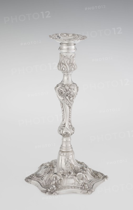 Single-candle candlestick by William Cafe fl. 1757–ca.1802, goldsmith. Rococo/Georgian George II style, 1763–1764, London, England, Great Britain. Raised domed foot with hexagonal rim decorated with flower heads at the corners, passing into a baluster shaft and terminating in a vase-shaped socket with a disc-shaped drippan. Decorated in rocaille motifs—plant tendrils, flowers, cartouches, shells, acanthus leaves—in slight spiral twist. Materials/technique: silver goldsmith work. Function: lighting. Height 29 cm; diameter 16.9 cm; weight 695 g., A tall ornate metallic candlestick stands upright against a pale gray background, showing a polished silver surface with decorative reliefs. The top has a shallow scalloped dish with floral and scroll motifs, leading to a slender stem that features raised acanthus-like leaves and rounded fluted sections. The midsection bulges into a heart-shaped knop decorated with sculpted roses and other floral forms, then tapers into a column with carved ridg...
