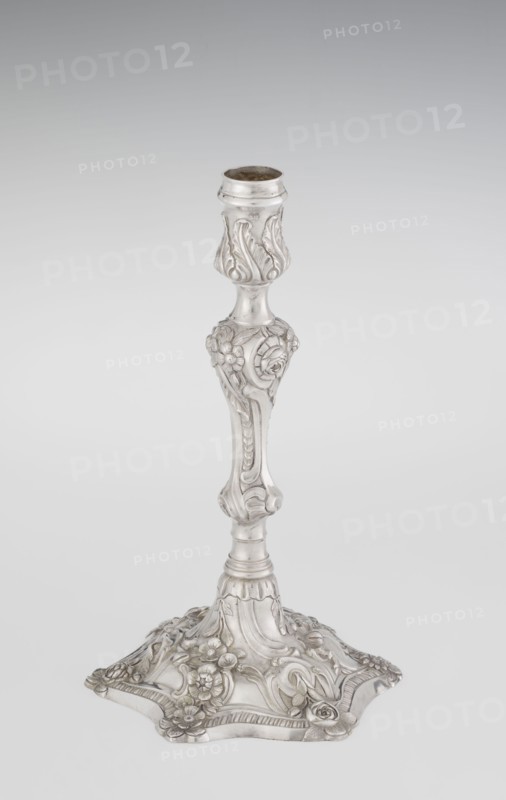 Single-candle candlestick by William Cafe fl. 1757–ca. 1802. Rococo/Georgian George II style, 1763/1764. Silver, single-candle candlestick on a raised, domed foot with a hexagonal rim, decorated with flower heads at the corners, passing into a baluster shaft and terminating in a vase-shaped socket with a disc-shaped drip pan. Ornament includes rocailles, plant tendrils, flowers, cartouches, shells, acanthus leaves and spiral-twisted relief decoration. Height 29 cm; diameter 16.9 cm; weight 695 g. Origin: London, England., A single ornate metal candlestick stands against a pale gray background, displaying a silvery finish with subtle darker tarnished areas; the upper socket has a flared rim and a bulbous section with raised leafy motifs, the slender stem features sculpted floral and scroll details and a few rounded nodes, and the broad base is shaped with curved edges and adorned with several raised flowers, leaves, and flowing decorative patterns in matching silver tones.