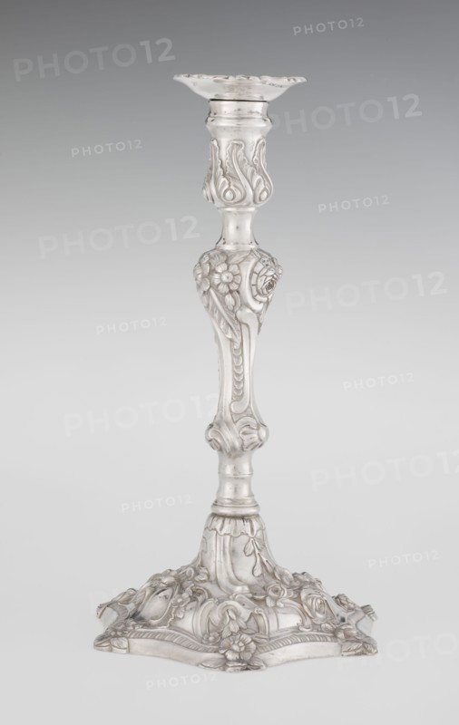 Single-candle candlestick by William Cafe fl. 1757–ca. 1802, rococo style, 1763/1764, London, England. Silver scrap construction, rococo on a raised, domed foot with a hexagonal rim decorated with flower heads at the corners, passing into a baluster shaft, terminating in a vase-shaped socket with a disc-shaped drip pan. Decorated with rococo motifs in the George II manner—rocailles, plant tendrils, flowers, cartouches, shells and acanthus leaves; the entire decoration is slightly spirally twisted. Function: lighting. Height 29 cm; diameter 16.9 cm; weight 717 g., A tall ornate candlestick made of a reflective silvery material stands against a smooth gray background, with a flared scalloped top, an elaborately carved central shaft featuring floral and leaf motifs, and a wide decorative base adorned with raised flowers, scrollwork, and curved panels; the visible colors are silver and shades of gray.