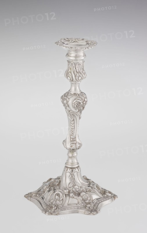 Single-candle candlestick, William Cafe fl. 1757–ca. 1802, goldsmith; rococo style, Georgian/George II influence; 1763–1764, London, England. Silver scrap-purchase technique. Rococo on a raised domed foot with hexagonal rim, decorated with flower heads at the corners, passing into a baluster shaft and terminating in a vase-shaped socket with a disc-shaped drip pan. Decorated with rocailles, plant tendrils, flowers, cartouches, shells and acanthus leaves; entire decoration slightly spirally twisted. Function: lighting. Height 29 cm; diameter 16.9 cm; weight 717 g., A single ornate candlestick stands against a light gray background, rendered in a silvery metallic tone. The piece has a flared, scalloped top with flowing carved edges, a slender central column with bulbous and contoured sections, and decorative floral and foliate reliefs along the shaft. The base is wide and irregularly shaped with raised floral motifs and curved, embossed patterns. The visible colors are silver and subtle ...