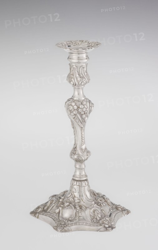 Single-candle candlestick, maker: William Cafe fl. 1757–ca. 1802, rococo style, 1763/1764, England — London, Great Britain. Single-candle candlestick on a raised, domed foot with a hexagonal rim, decorated with flower heads at the corners, passing into a baluster shaft and terminating in a vase-shaped socket with a disc-shaped profilm profit. Candlestick decorated with rococo motifs in the George II style — rocailles, plant tendrils, flowers, cartouches, shells and acanthus leaves; the entire decoration is slightly spirally twisted. Materials/technique: scrap goldsmith. Function: lighting. Dimensions: height 29 cm, diameter 16.9 cm, weight 717 g., A silver-colored ornate candlestick with a decorative round top and a flared, scalloped rim, a tapered shaft featuring raised floral and leaf motifs, a bulbous midsection with embossed flowers and scrolling details, and a wide decorative base with embossed flowers, leaves, and curved ornamental patterns, all shown against a pale gray to white...
