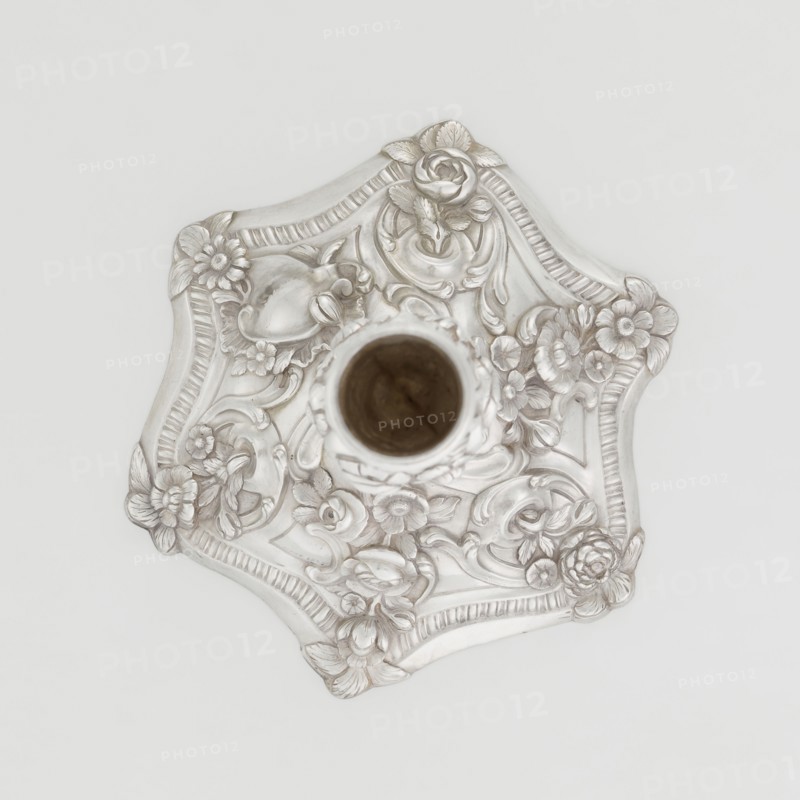 Single-candle candlestick by William Café fl. 1757–ca. 1802, rococo, 1763–1764, England, London. Rococo single-candle candlestick on a raised domed foot with a hexagonal rim, decorated with flower heads at the corners, passing into a baluster shaft and terminating in a vase-shaped socket with a disc-shaped drip pan. Decorated with rococo motifs in the George II style — rocailles, plant tendrils, flowers, cartouches, shells and acanthus leaves — with the entire decoration slightly spirally twisted. Materials/technique: scrap. Function: lighting. Dimensions: height 29 cm, diameter 16.9 cm, weight 717 g., A decorative metallic object with an ornate floral relief seen from above, featuring a central cylindrical opening and a scalloped edge; the surface shows raised sculpted flowers, leaves, and scrollwork in a silvery finish with subtle darker shadows and a small area of brown visible inside the central opening, all set against a pale off-white background.