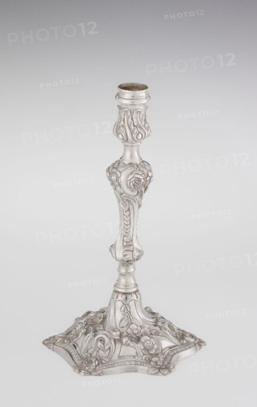 Single-candle candlestick, William Cafe fl. 1757–ca.1802, rococo George II style. Silver scrap—single-candle candlestick on a raised, domed foot with a hexagonal rim decorated with flower heads at the corners, passing into a baluster shaft and terminating in a vase-shaped socket with a disc-shaped drip pan. Decorated with rococo motifs: rocailles, plant tendrils, flowers, cartouches, shells and acanthus leaves; overall decoration slightly spirally twisted. Dated 1763/1764. London, England. Lighting. Height 29 cm; diameter 16.9 cm; weight 717 g., A single ornate candlestick stands against a plain light gray background; the candlestick is metallic silver in color with a polished sheen and features intricate carved floral and leaf motifs along its shaft and base, including rounded shapes, scrolling patterns, and raised rose-like flowers; the base spreads outward into a decorative, scalloped form with more floral relief and curved accents, and the top has a cylindrical holder with a slight...