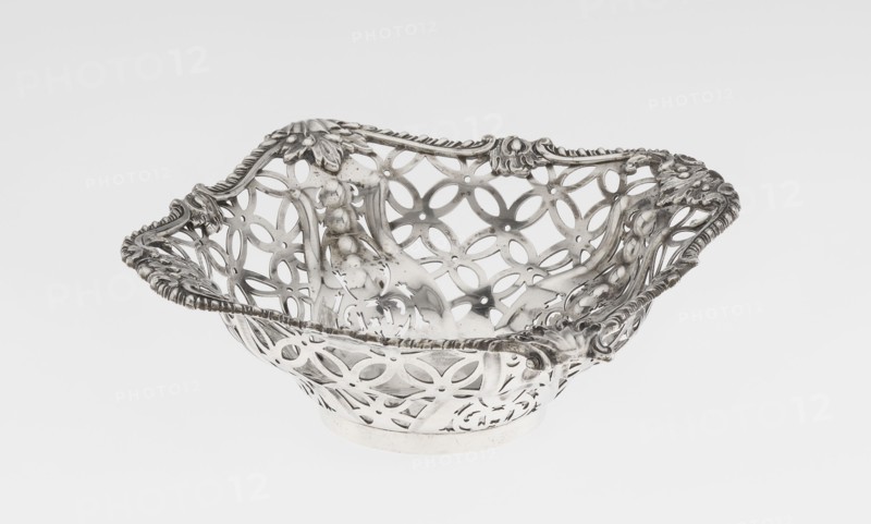Openwork basket by Comyns & Sons goldsmiths’ firm, active 1859–1953. Four-sided openwork basket on a circular foot, decorated with four-leaf rosette motifs, smooth stylized leaves and ribbons in the corners; irregularly cut edge enclosed in a godron half-cylinder and decorated with acanthus leaves. Materials/technique: openwork, godron ornaments. Date: 1904–1905. Place: London, England, Great Britain. Function: vessel. Dimensions: height 4.5 cm; width 15 cm; depth 15 cm; weight 155 g., A decorative metallic bowl with an openwork floral and geometric pattern, showing ornate raised edges with scrolling leaf and rope-like details; the bowl has a reflective silver surface with areas of darker tarnish and subtle shine, and the background is a smooth pale gray to white gradient.