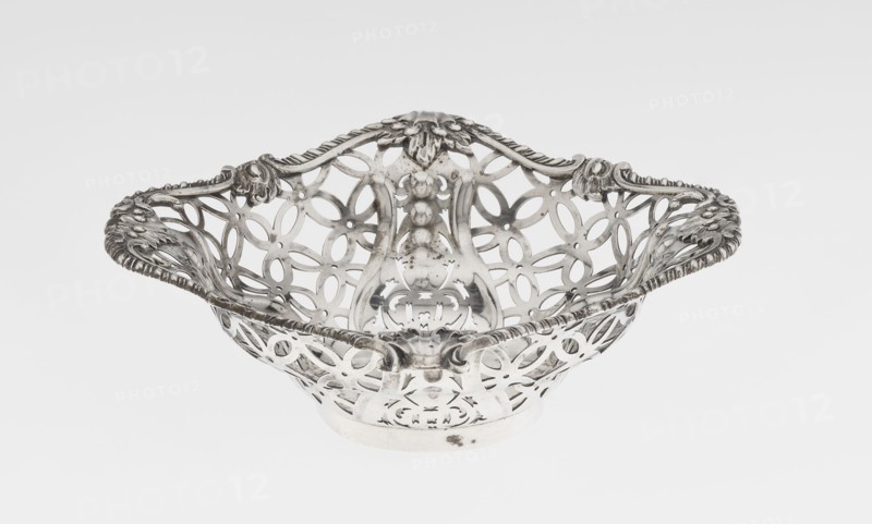 Openwork basket by Comyns & Sons London; goldsmiths’ firm, fl. 1859–1953. Basket on a circular foot, four-sided openwork, decorated with a motif of four-leaf rosettes and smooth stylised leaves and ribbons in the corners; irregularly cut edge enclosed in a godron half-cylinder and decorated with acanthus leaves. Materials/technique: openwork metal with godron and acanthus ornament. Date: 1904/1905. Place: London, England, Great Britain. Classification: vessel. Dimensions: height 4.5 cm; width 15 cm; depth 15 cm; weight 155 g., A decorative metal basket with an openwork lattice pattern and ornate scrolling edges, shown against a plain light background. The basket has a shallow bowl shape with curved handles integrated into the rim and a raised central section with embossed floral and bead-like motifs. The metal surface shows polished and slightly tarnished areas. Visible colors include silver-gray tones and subtle darker gray spots.