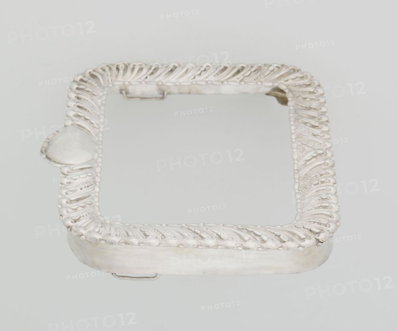 Dressing mirror frame, Comyns & Sons London; goldsmiths; fl. 1859–1953 maker, scrap, clasp, medallion; rectangular mirror frame with rounded corners decorated with oblique clasp and oval medallion; 1890–1891; Great Britain, England, London; dressing accessories; frame; height 15.7 cm, width 13.2 cm, depth 2 cm, weight 102 g, A square decorative frame with rounded corners, shown against a pale background, featuring an ornate raised border with a repeating curved leaf-like pattern and small bead-like accents; the frame appears metallic with a shiny silver surface and some areas of subtle darker tarnish, and there is a small rounded protrusion on one side and a few clip-like fixtures along the inner edge.