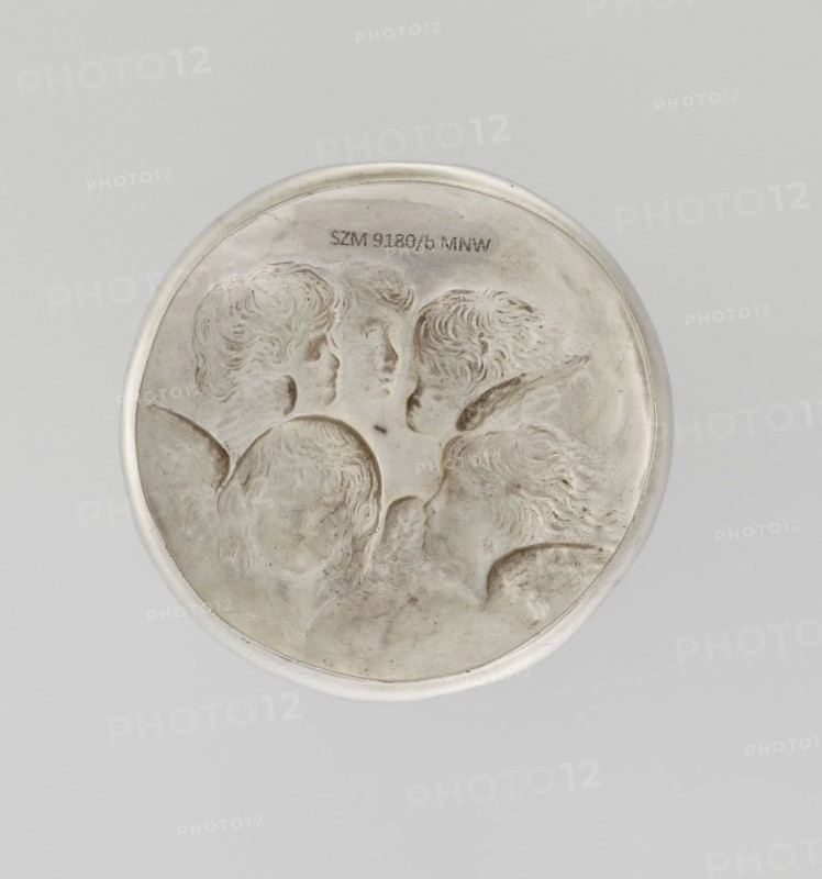 Toilet box, Comyns & Sons London; goldsmiths, fl. 1859–1953. Cylindrical toilet box, decorated on the mantle with a relief plant braid forming four reserves containing winged angel heads in clouds; raised, flattened cover decorated with four putti heads. Depictions after a painting by Joshua Reynolds Angel's Heads, 1786. 1898–1899. England, London. Materials/subject: putti, putti heads, angel iconography, plant ornaments. Function: toilet accessories. Height 9.2 cm; diameter 7.5 cm; weight 147 g., A round metallic object with a raised rim, showing a relief of a group of several heads with carved hair and facial profiles, rendered in shades of silver and pale gray with some darker gray areas and subtle brownish discoloration, set against a light gray background.