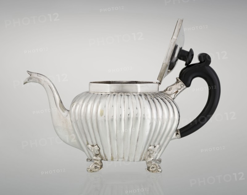 Teapot, maker Thomas II fl. 1798–1809?, goldsmith. Teapot supported on four legs in the form of dolphins, octagonal, decorated with vertical narrow ringlets and flutes, with a spout-shaped spout, with a wooden handle not original mounted in sockets, with a slightly arched hinged lid, wooden finial/handle not original. Iconography: dolphin; function: tea vessel. Date: 1808–1809. Place: London, England, Great Britain. Materials: silver implied by goldsmith and wood handle. Dimensions: height 12 cm, width 20.5 cm, depth 14 cm; weight 595 g. Provenance note: scrap - purchase; purchase., A silver teapot with a fluted, rounded body standing on a group of ornate feet, a curved spout on one side and a black curved handle on the other, the hinged lid is open showing the inner rim; visible colors include silver, black, and subtle reflections of gray.