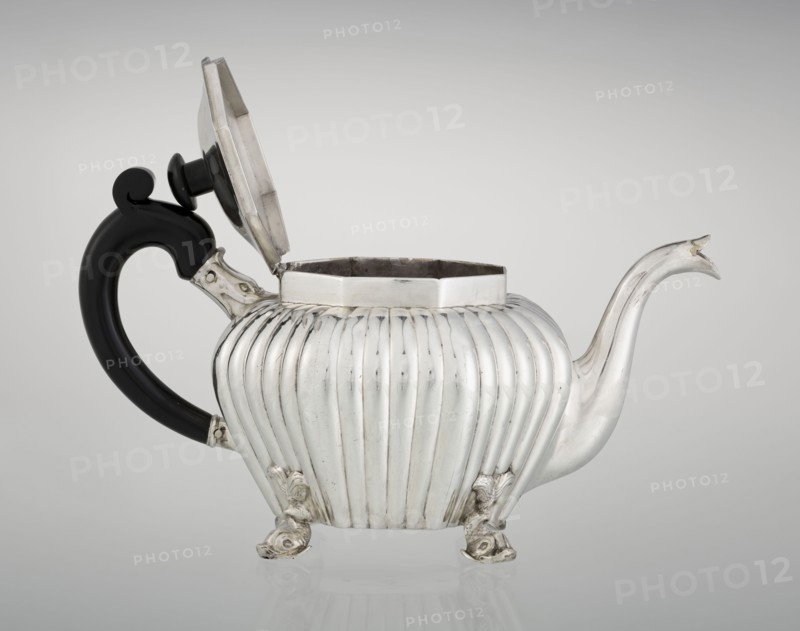 Teapot, maker Thomas II fl. 1798–1809?, goldsmith. Teapot supported on four legs in the form of dolphins; octagonal body decorated with vertical narrow ringlets and flutes; spout-shaped spout; slightly arched hinged lid with wooden handle not original; additional wooden handle not original mounted in sockets. Iconography: dolphin; function: tea vessel. Date: 1808/1809. Place: London, England, Great Britain., silver scrap?, tea vessel. Dimensions: height 12 cm, width 20.5 cm, depth 14 cm; weight 595 g. Provenance: purchase., A silver teapot with a ribbed, rounded body sits on a reflective surface, its hinged lid open and revealing a dark interior; the teapot has a curved spout with a pointed tip and a black, curved handle attached with silver fittings, and it rests on decorative feet shaped like stylized animal forms. Colors visible include silver, black, dark gray, and pale gray in the background.