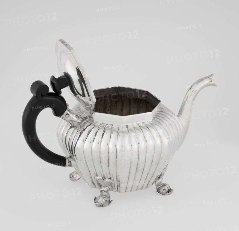 Teapot, Holland, Thomas II fl. 1798–1809? goldsmith, scrap - purchase, tea vessel, dolphin iconography, ringing, tea vessels. Teapot supported on four legs in the form of dolphins, octagonal, decorated with vertical narrow ringlets and flutes, with a spout-shaped spout, with a wooden handle not original mounted in sockets, with a slightly arched hinged lid, with a wooden handle not original. 1808/1809. England, London. Vessel. Height 12 cm; width 20.5 cm; depth 14 cm; weight 595 g., A metallic teapot with a ribbed, rounded body and a hinged lid is shown with the lid open, revealing a dark interior; the teapot has a curved spout and rests on a group of ornate feet, and it features a black, curved handle with a decorative knob at the top; visible colors include silver, black, and shades of gray.