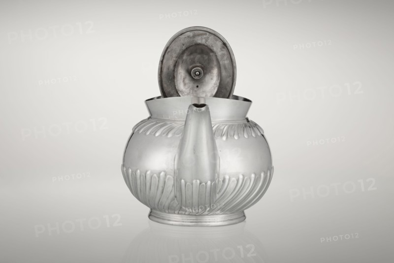 Teapot, made by Goldsmiths & Silversmiths Co Ltd London; makers William Gibson & John Langman goldsmiths. Egg-shaped teapot on an oval foot, decorated with two bands of diagonal clasp, with a high upper edge, hinged lid topped with a handle decorated with a wooden rosette, prominent spout on the side, non-original wooden handle set in silver sockets. Tea vessel tea accessory. Made 1896/1897, Sheffield, England. Dish. Height 13.5 cm, width 22.8 cm, depth 13.8 cm, weight 666 g., A metallic teapot with a rounded body and an open hinged lid is centered against a light gray background, showing a smooth reflective surface with decorative vertical ridges around its lower and upper halves, a short rounded spout at the front, and a slightly raised circular base; the visible colors are silver, gray, and subtle darker gray tones inside the lid.