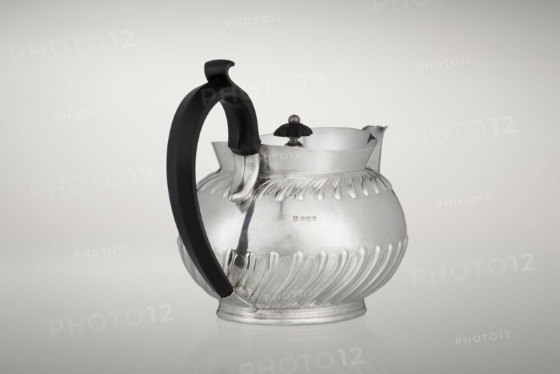 Teapot by Teapot, Goldsmiths & Silversmiths Co Ltd; makers William Gibson & John Langman goldsmiths. Egg-shaped silver teapot on an oval foot, decorated with two bands of diagonal clasp, high upper edge, hinged lid topped with a handle decorated with a wooden rosette, prominent spout, non-original wooden handle set in sockets. Made in modern times, 1896–1897, Sheffield, England. Height 13.5 cm; width 22.8 cm; depth 13.8 cm; weight 666 g., A rounded metal pitcher with a reflective silver surface and decorative fluted ridges around its lower and upper body sits against a light neutral background, featuring a curved black handle attached to one side and a scalloped spout on the opposite side, with a small dark knob on a short stem at the top; visible colors include silver, black, and shades of gray, and the pitcher rests on a slightly raised circular base with its reflection faintly visible on the surface beneath.