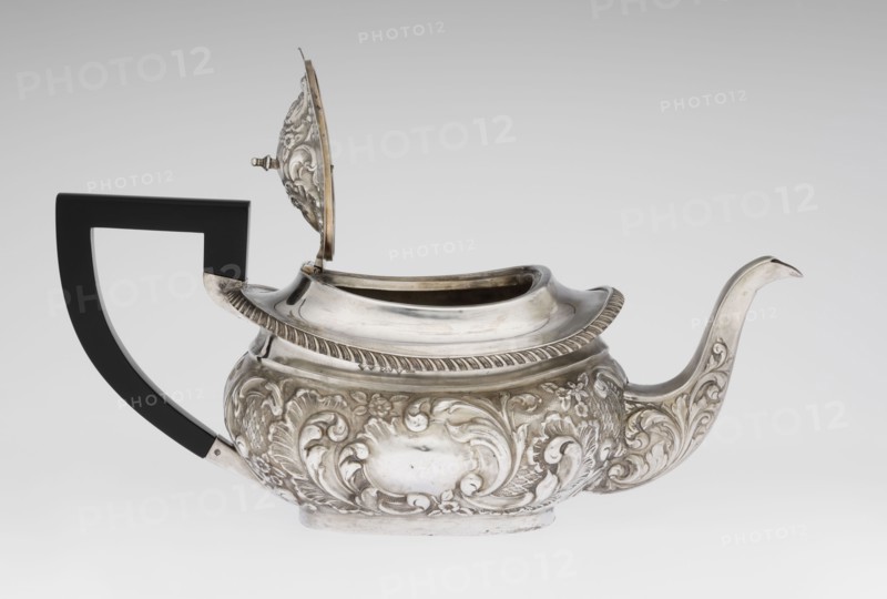 Teapot by Henry Moreton fl. 1899–1930s, goldsmith and factory owner; neo-Rococo style, relief decoration, acanthus leaves and godrons ornamentation. Egg-shaped teapot on an oval plinth with a side spout, sleeves for attaching a wooden handle mounted on the axis handle added contemporarily. Upper edge bordered with a band of godrons and a wide collar; hinged, centrally arched lid topped with a handle. Oval blind reserve on the belly with acanthus rim. Dated 1909/1910, made in Chester, England. Vessel; height 13.5 cm, width 26 cm, depth 12 cm, weight 555 g., A silver teapot with ornate raised floral and scrollwork decoration across its rounded body and spout, a hinged lid that is open showing the interior rim, and a curved spout with decorative motifs; the teapot has a contrasting angular black handle attached at the back, a ribbed edge around the top, and a slightly raised base. The visible colors are silver, black, and subtle variations of gray on the background and reflected surfaces.