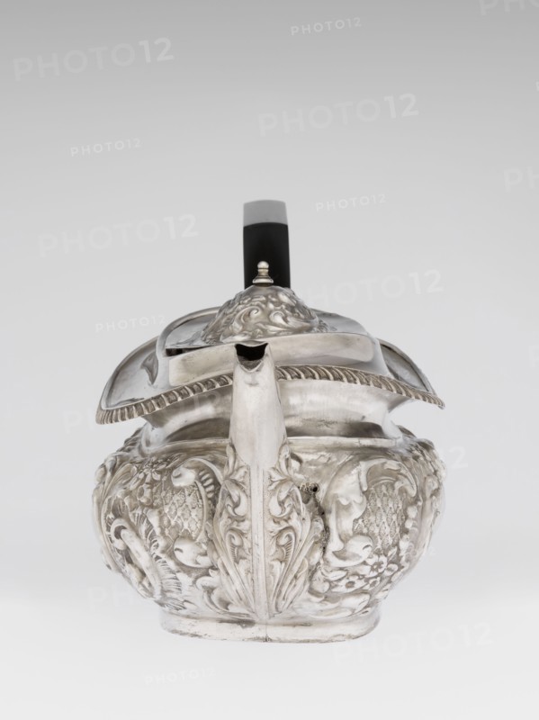Teapot by Henry Moreton fl. 1899–1930s, factory owner and goldsmith; neo-Rococo style; silver; 1909/1910, Chester, England. Egg-shaped teapot on an oval plinth with a side spout, sleeves on the axis for a wooden handle added contemporarily, hinged centrally arched lid topped with a handle. Upper edge bordered with a band of godrons and a wide collar. Entire surface decorated in neo-Rococo relief motifs and acanthus leaves, with an oval blind reserve on the belly framed by an acanthus rim. Function: tea vessel. Height 13.5 cm; width 26 cm; depth 12 cm; weight 555 g., A silver teapot with ornate embossed floral and scrollwork decoration sits against a gray to white gradient background; the body is rounded with raised decorative panels and a sculpted vertical ridge at the front that integrates into the short spout, the hinged lid has a domed finial and a scalloped edge, and a black handle with a gray top extends upward from the back; visible colors include silver, black, gray, and white.