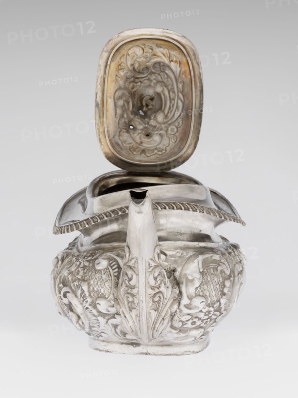 Teapot by Henry Moreton fl. 1899–1930s, factory owner, goldsmith; neo-Rococo style, relief decoration. Egg-shaped teapot on an oval plinth with spout-shaped side spout; sleeves for attaching a wooden handle mounted on the axis wooden handle added contemporaneously. Upper edge bordered by a band of godrons with a wide collar above; hinged, centrally arched lid topped with a handle. Decorated overall with neo-Rococo relief motifs and acanthus leaves; oval blind reserve on the belly with acanthus rim. Dated 1909/1910. England, Chester. Vessel. Height 13.5 cm, width 26 cm, depth 12 cm, weight 555 g., A sculpted metal vessel with an open hinged lid sits against a light gray background; the vessel has a rounded body with ornate relief decoration featuring flowing leaf and floral motifs and textured areas, and a prominent spout rising from the front that is decorated with a vertical leaf-like design. The hinged lid has an inner surface with carved swirling patterns and a patina that shows war...