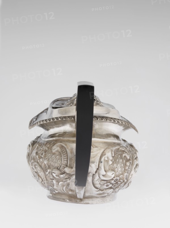 Teapot by Henry Moreton fl. 1899–1930s, factory owner and goldsmith; neo-Rococo style; silver; relief decoration with neo-Rococo motifs and acanthus leaves; teapot on an oval plinth, egg-shaped body with spout on the side; sleeves for attaching a wooden handle mounted on the axis added contemporarily; upper edge bordered with a band of godrons and a wide collar; hinged, centrally arched lid topped with a handle; oval blind reserve on the belly with acanthus rim; dated 1909/1910; Chester, England; vessel. Dimensions: height 13.5 cm, width 26 cm, depth 12 cm; weight 555 g., An ornate metal vessel with a rounded body and embossed decorative motifs sits centered in the image, viewed from the front with a tall straight handle rising from its top to bottom; the vessel surface shows intricate raised floral and scroll patterns and a scalloped rim, and the handle appears smooth and solid. The visible colors are silver on the metal surface and black on the handle, set against a pale gray to whit...