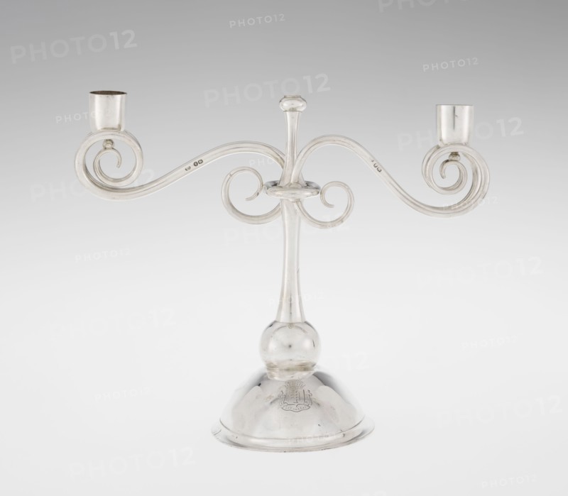 Two-candle candlestick. Maker: William Leuchars fl. 1876–1888; firm: Leuchars & Son London, goldsmiths, fl. ca. 1845–1902., silver lighting fixture, two-branched candlestick on a circular domed foot. Description: shaft rests on a ball no finial, enclosed at 3/4 height in a plate-like ring from which extend movable arms of S-shaped twisted wires, split at the ends, terminating in cylindrical candle sockets. Foot engraved with the Pilawa coat of arms with motto beneath a nine-baton count's crown; includes coats of arms and heraldic rank crowns. Date: 1881–1882. Place of origin: London, England, Great Britain. Dimensions: height 27 cm; width 32 cm; depth 11.5 cm; weight 1082 g., A silver-colored metal candelabrum with a central vertical stem rising from a rounded base, a bulbous knop near the lower stem, and two symmetrically curved arms that curl into spiral scrolls ending in cylindrical candle cups; the surface is reflective and shows highlights and subtle darker tarnish on some areas, ...