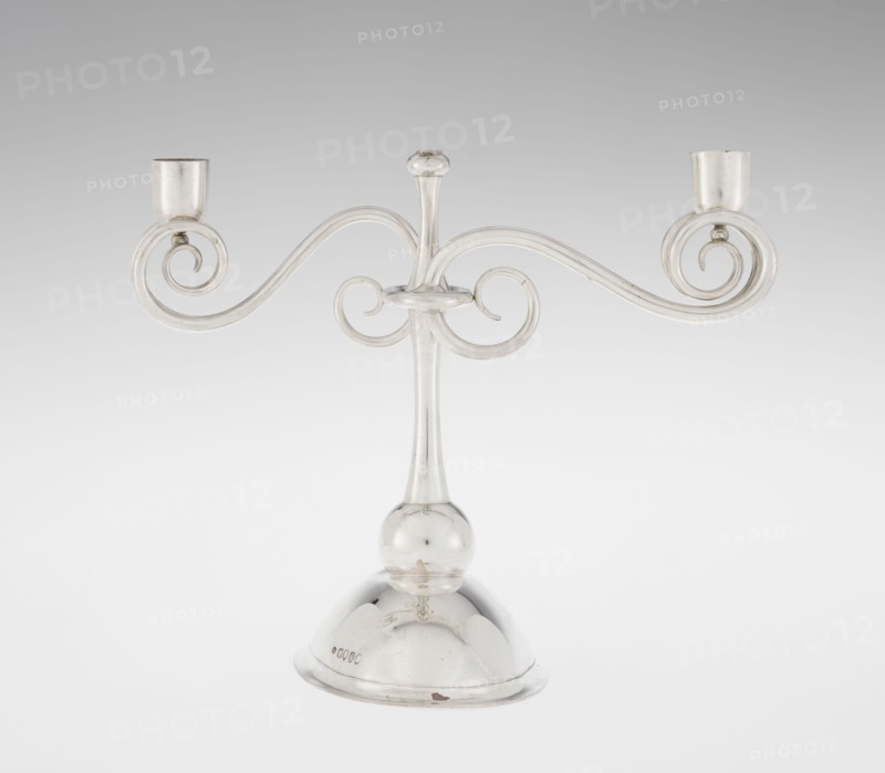 Two-candle candlestick by William Leuchars fl. 1876–1888, Leuchars & Son London, goldsmith's firm, fl. ca. 1845–1902. Two-branched candlestick on a circular domed foot. The shaft rests on a ball no finial and is enclosed at three-quarters of its height by a plate-like ring, from which extend movable arms made of S-shaped twisted wires, split at the ends and terminating in cylindrical candle sockets. The foot is engraved with the Pilawa coat of arms and motto beneath a nine-baton count's crown. Date: 1881/1882. Origin: London, England. Function: lighting. Dimensions: height 27 cm; width 32 cm; depth 11.5 cm. Weight: 1082 g., A metallic candlestick with a polished silver appearance stands on a rounded dome base and a spherical joint, rising into a slender central stem topped with a small rounded finial; two ornate horizontal arms extend from the stem in symmetrical curved scrolls ending in cup holders, creating an overall elegant, curving silhouette. The visible colors are silver and sha...