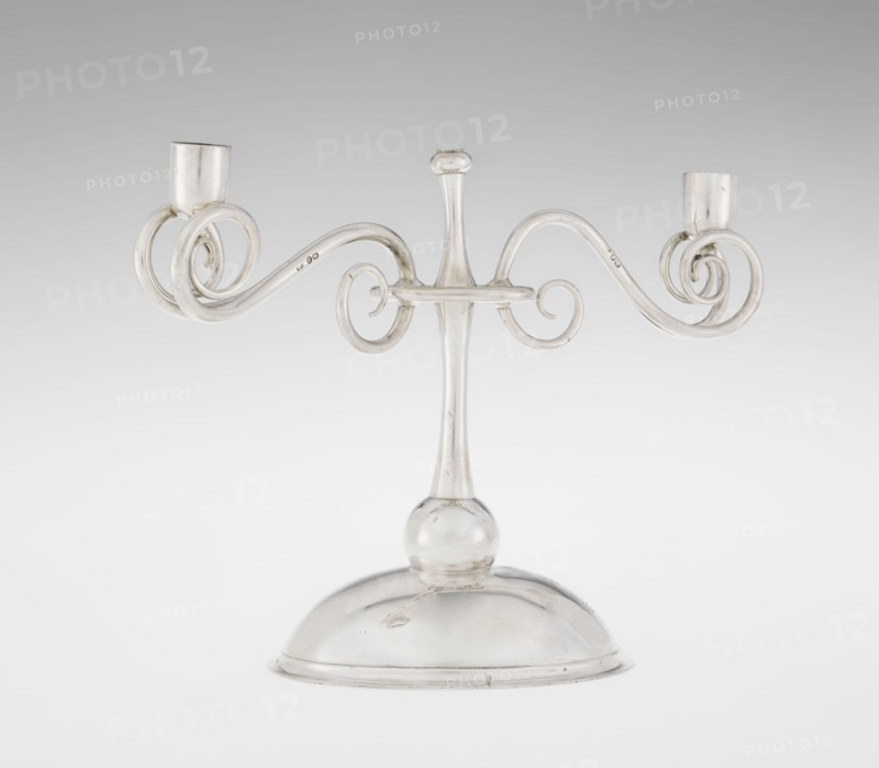 Two-candle candlestick, attributed to William Leuchars fl. 1876–1888 and the firm Leuchars & Son London; goldsmiths, fl. ca. 1845–1902. Silver two-branched candlestick on a circular domed foot. The shaft rests on a ball no finial and is enclosed at three-quarters of its height by a plate-like ring, from which extend movable arms made of S-shaped twisted wires, split at the ends and terminating in cylindrical candle sockets. The foot is engraved with the Pilawa coat of arms beneath a nine-baton count’s crown, with mottos. Date: 1881–1882. Place of origin: London, England, Great Britain. Function: lighting. Dimensions: height 27 cm; width 32 cm; depth 11.5 cm; weight 1082 g., A clear glass two-armed candlestick with a rounded dome base and a slender central stem topped by a small bulb, featuring ornate curved scroll arms that extend outward and curl into circular spirals ending in short cylindrical holders; the visible colors are clear glass with subtle highlights of white and pale gray ...